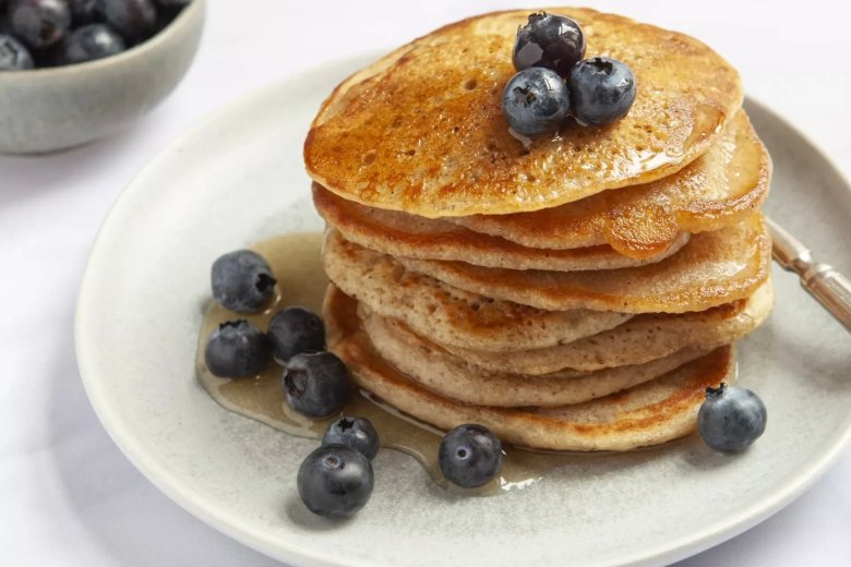 A stack of American-style pancakes topped with blueberries and drizzled with syrup sits on a plate, reminiscent of a mouthwatering recipe from the Hairy Bikers' Chicken &amp; Egg series. A bowl of fresh blueberries awaits beside it.