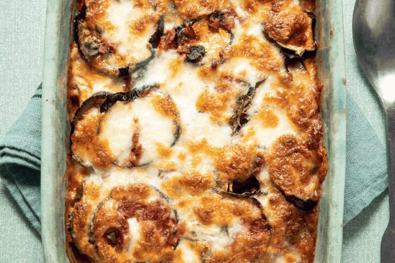 A baked casserole dish of golden-brown, cheesy Aubergine Parmigiana inspired by the Hairy Dieters’ Simple Healthy Food sits on a folded green cloth next to a metal spoon, viewed from above.