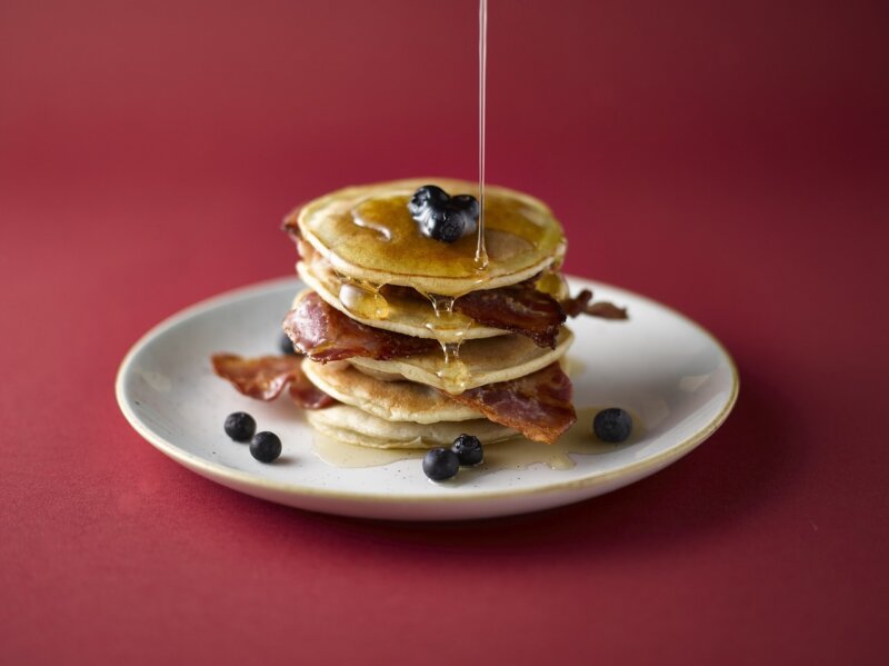 A stack of pancakes topped with blueberries and crispy bacon, drizzled with syrup, served on a white plate against a red background, reminiscent of the recipes in Hairy Bikers 'Mum's Know Best' cookery book.