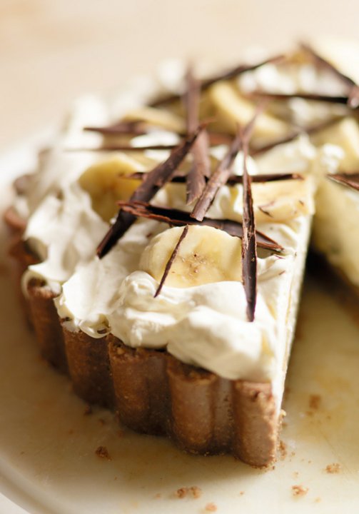 A close-up of a banana cream pie inspired by the Hairy Bikers, topped with whipped cream, banana slices, and decorative shards of dark chocolate, with one slice partially removed.