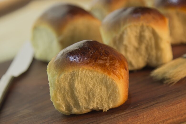 Close-up view of several basic golden brown bread rolls on a wooden surface, accompanied by a butter knife and a basting brush.