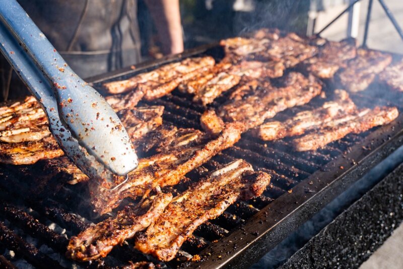 A pair of metal tongs holds a piece of seasoned meat over an open grill loaded with multiple BBQ meats, all being barbecued.