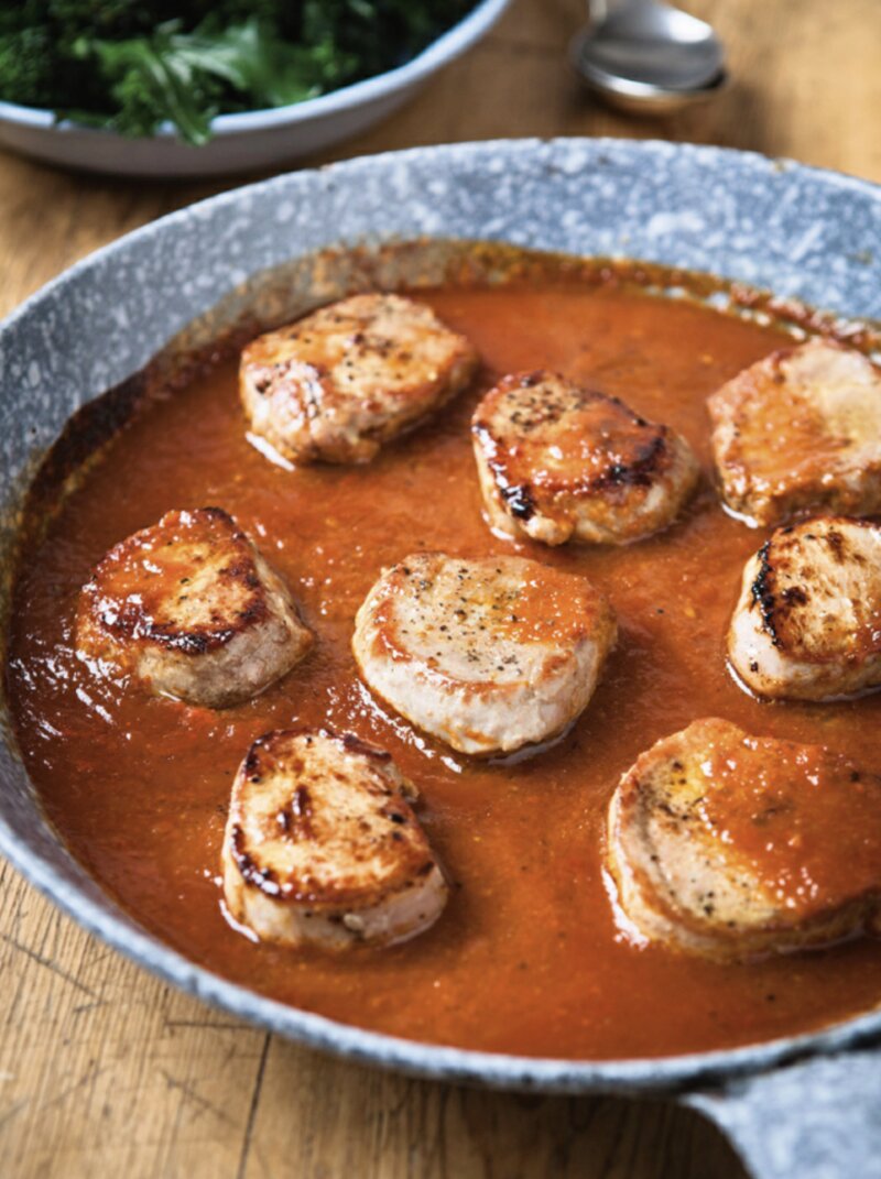 Sliced pork medallions in BBQ sauce are seared and simmering in a tomato-based mixture in a frying pan, with a plate of greens in the background, offering a fast food twist on this savoury dish.
