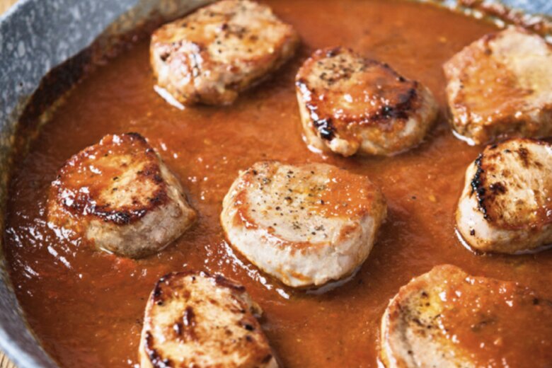 Sliced pork medallions in BBQ sauce are seared and simmering in a tomato-based mixture in a frying pan, with a plate of greens in the background, offering a fast food twist on this savoury dish.