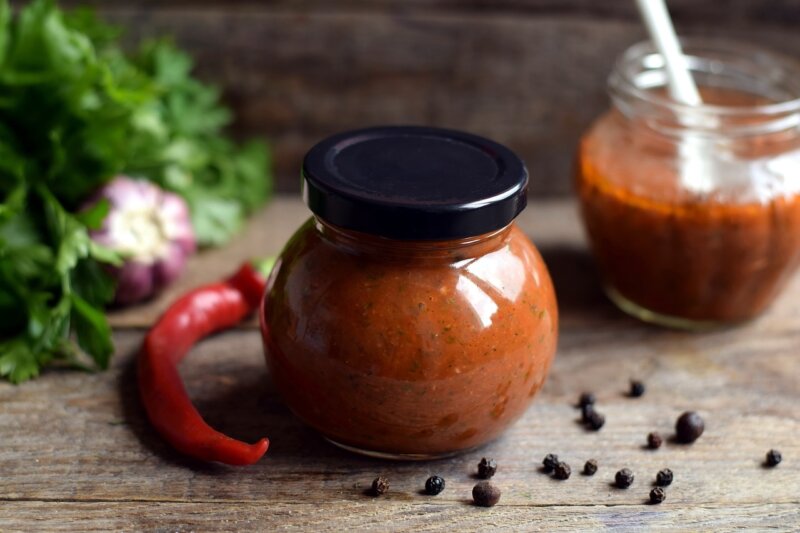 Two glass jars with homemade BBQ sauce, one closed with a black lid and the other open with a spoon. A red chili, garlic, black peppercorns, and fresh herbs are strewn around on a wooden surface — perfect companions for Kingies homemade BBQ recipes.