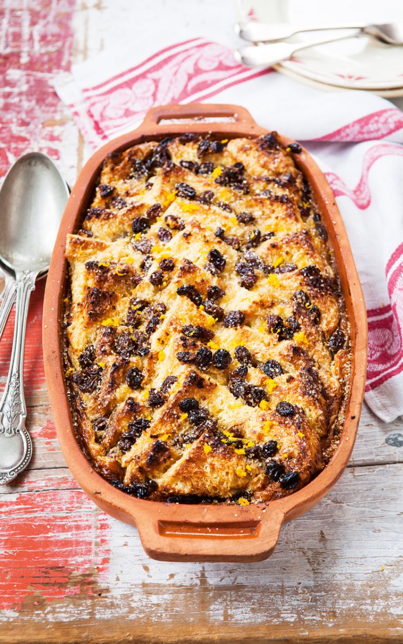 A classic Bread and Butter Pudding topped with sultanas is served in a rectangular ceramic dish on a rustic wooden table, reminiscent of the comforting dishes from Hairy Bikers Mediterranean Adventure, with a red and white cloth and silver spoons beside it.