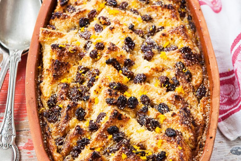 A classic Bread and Butter Pudding topped with sultanas is served in a rectangular ceramic dish on a rustic wooden table, reminiscent of the comforting dishes from Hairy Bikers Mediterranean Adventure, with a red and white cloth and silver spoons beside it.