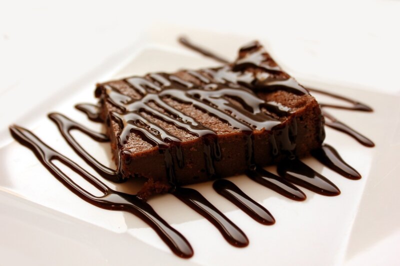 A square piece of chocolate brownie drizzled with chocolate sauce on a white plate, reminiscent of decadent sweet potato brownies from the Hairy Bikers.