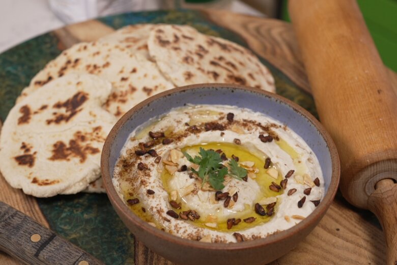 A bowl of butterbean hummus garnished with herbs, nuts, and olive oil, placed on a green plate with several pieces of grilled flatbreads and a rolling pin beside it.