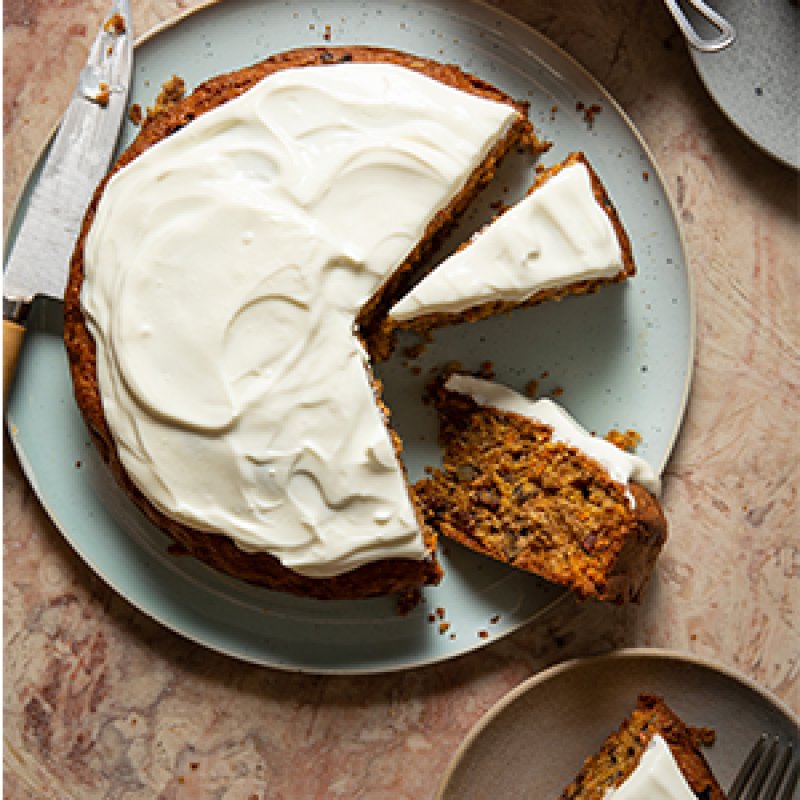 A round Carrot Cake with creamy white icing, partially sliced. One slice is served on a small plate, with a knife resting nearby on the main plate—a tempting example of simple healthy food on a light-coloured marble surface.