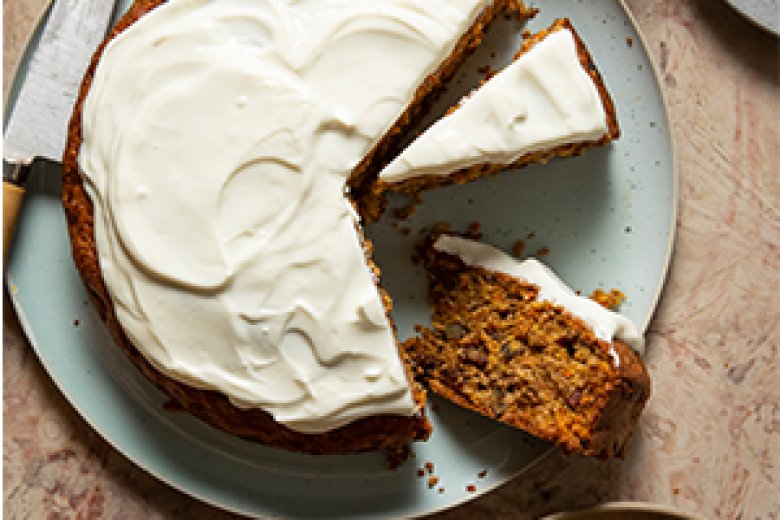 A round Carrot Cake with creamy white icing, partially sliced. One slice is served on a small plate, with a knife resting nearby on the main plate—a tempting example of simple healthy food on a light-coloured marble surface.