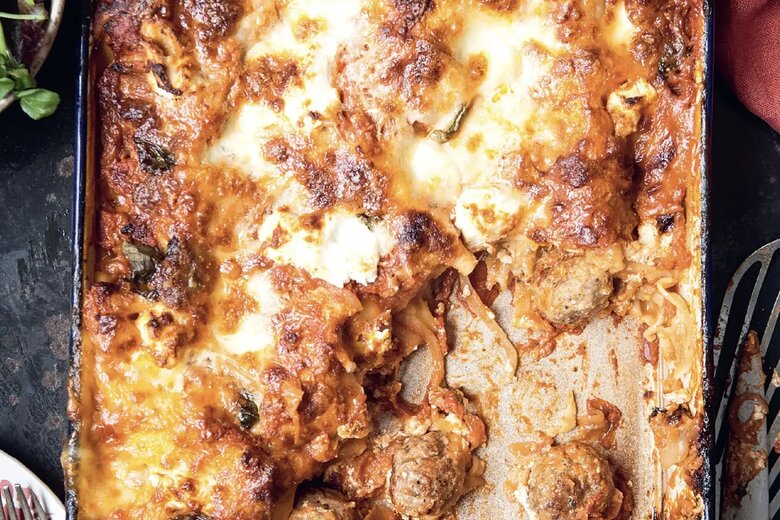 A celebratory lasagne sits in a rectangular pan with some portions removed, revealing layers of pasta, meatballs, sauce, and cheese. A plate with salad and cutlery rests beside the pan, reminiscent of the comfort found in the best of the Hairy Bikers' creations.