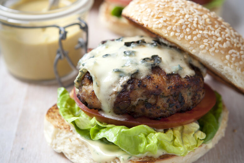 A close-up of a chicken burger from the Hairy Bikers 'chicken and egg' cookbook, with a sesame seed bun, lettuce, tomato, grilled patty topped with melted blue cheese, and a jar of creamy sauce in the background. Another burger is slightly out of focus behind it.