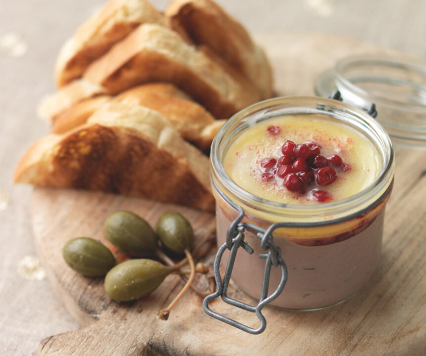A glass jar of Chicken Liver Parfait with Cranberry Butter, inspired by the Hairy Bikers - 12 Days of Christmas recipe, topped with cranberries and clarified butter, served on a wooden board with toasted bread and caperberries.