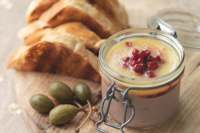 A glass jar of Chicken Liver Parfait with Cranberry Butter, inspired by the Hairy Bikers - 12 Days of Christmas recipe, topped with cranberries and clarified butter, served on a wooden board with toasted bread and caperberries.