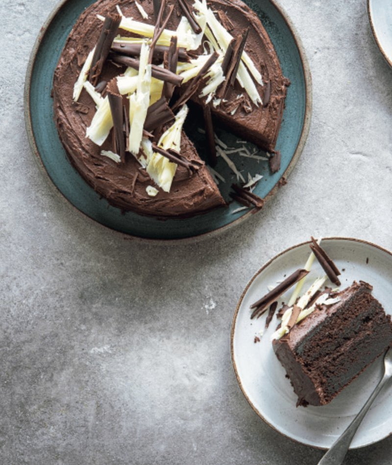A round Chocolate Cake topped with white and dark chocolate shavings, inspired by the Hairy Bikers' Everyday Winners, with one slice served on a separate plate.