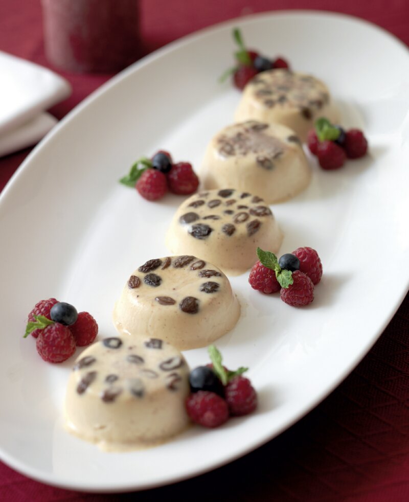 A white oval plate with five round Christmas panna cotta desserts topped with chocolate chips, garnished with fresh raspberries, blueberries, and mint leaves, set on a red tablecloth—perfect for the Hairy Bikers' 12 Days of Christmas cookery book.
