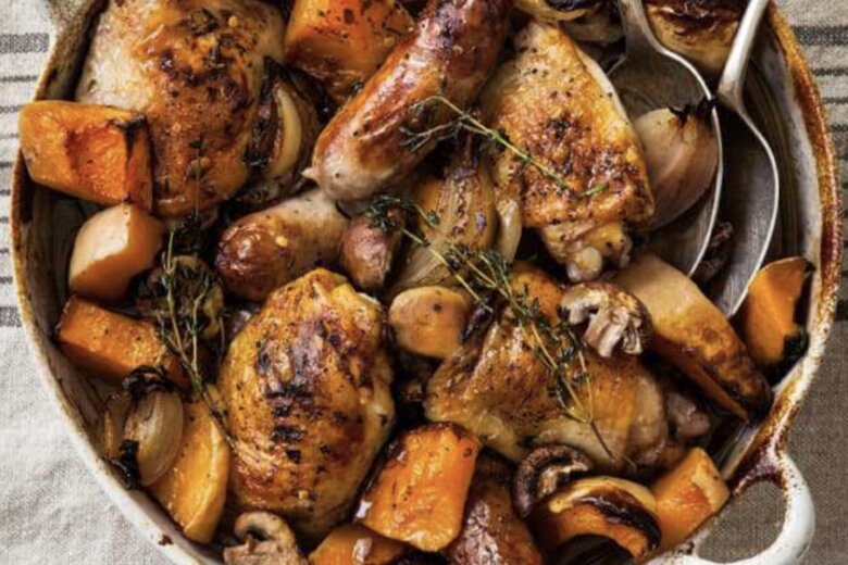 A rustic chicken and squash traybake inspired by British classics, with roast chicken, sausages, pumpkin, onions, and mushrooms garnished with herbs in a white round casserole dish on a striped cloth background.