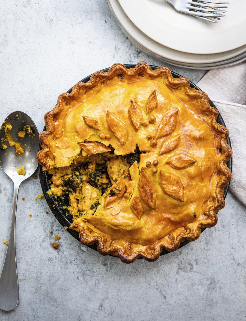 Golden-brown Curried Chicken Pie with decorative leaf-shaped pastry, partially sliced to reveal a savoury filling, sits on a grey surface beside a serving spoon - just like a recipe from the Hairy Bikers’ Brilliant Bakes cookbook.
