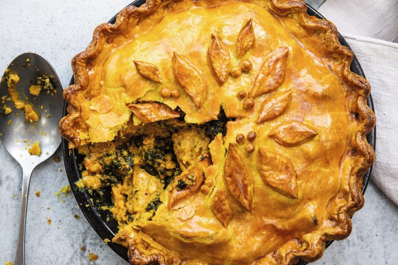 Golden-brown Curried Chicken Pie with decorative leaf-shaped pastry, partially sliced to reveal a savoury filling, sits on a grey surface beside a serving spoon - just like a recipe from the Hairy Bikers’ Brilliant Bakes cookbook.