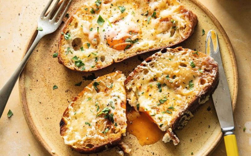 Toasted bread slices with melted cheese and herbs sit invitingly on a plate, reminiscent of family favourites. One slice is cut open, revealing egg yolk oozing perfectly, accompanied by a fork and knife—a dish the Hairy Bikers love.