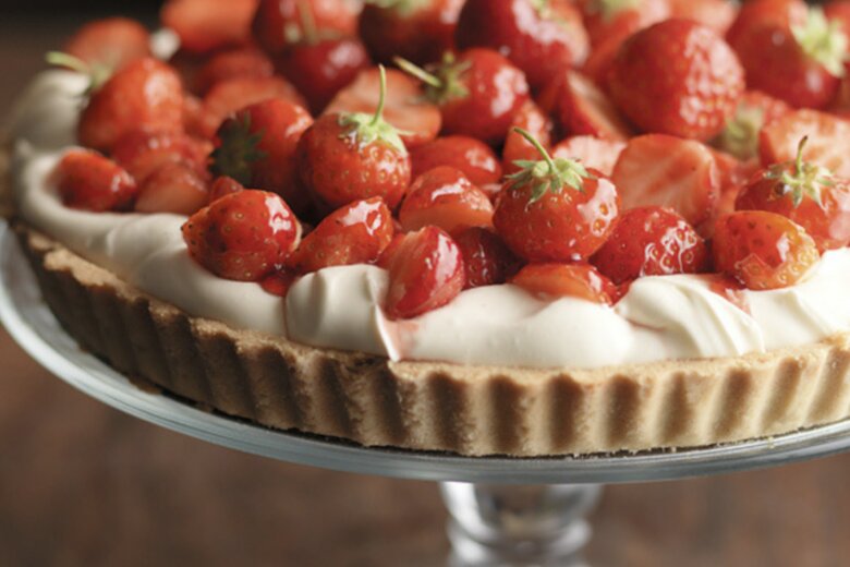 A strawberry tart topped with whole glazed strawberries sits on a glass cake stand, looking like it belongs in one of the Hairy Bikers' perfect pies recipes.