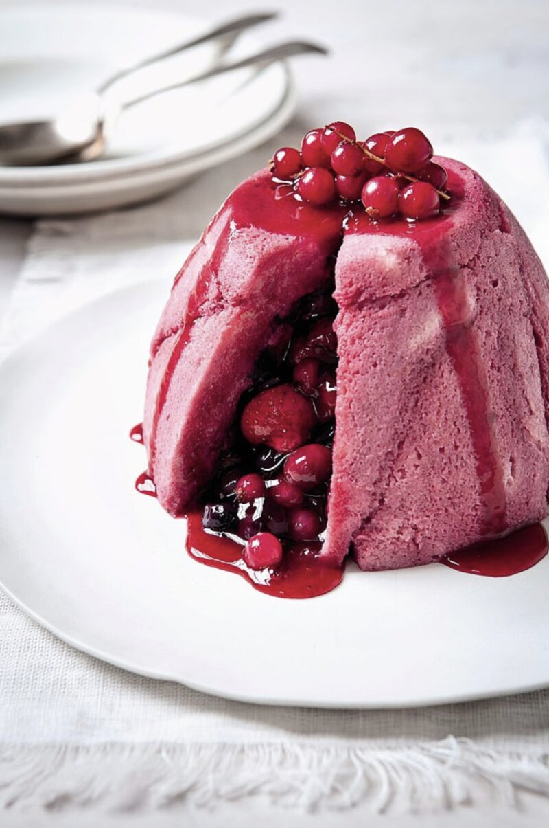 A delightful summer pudding with a pink sponge base and berry filling, garnished with fresh berries on top and rich berry sauce dripping down, is placed elegantly on a white plate. Forks and plates in the background hint at good eating to come.