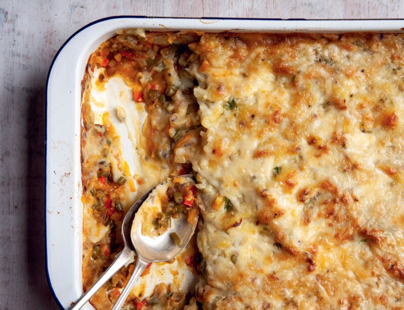 A baking dish with a partially served Indian Shepherds Pie, showing layers of spiced vegetables, and melted cheese, with two serving spoons resting inside—a cosy dish inspired by the Hairy Bikers' Veggie Feasts.