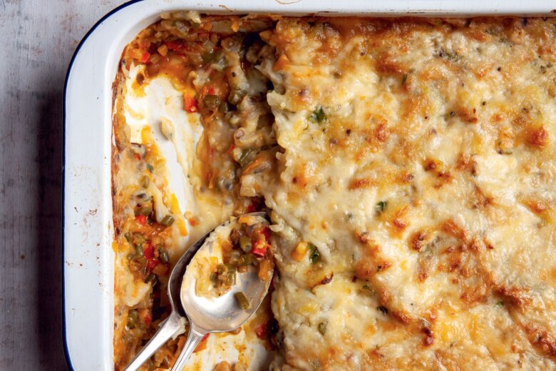 A baking dish with a partially served Indian Shepherds Pie, showing layers of spiced vegetables, and melted cheese, with two serving spoons resting inside—a cosy dish inspired by the Hairy Bikers' Veggie Feasts.