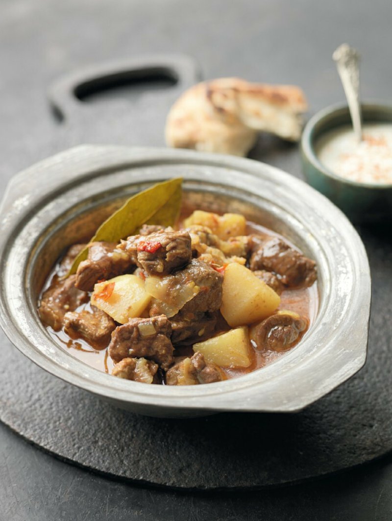 A bowl of Lamb Vindaloo with potatoes and bread on a table from the Hairy Bikers' Great Curries recipe book