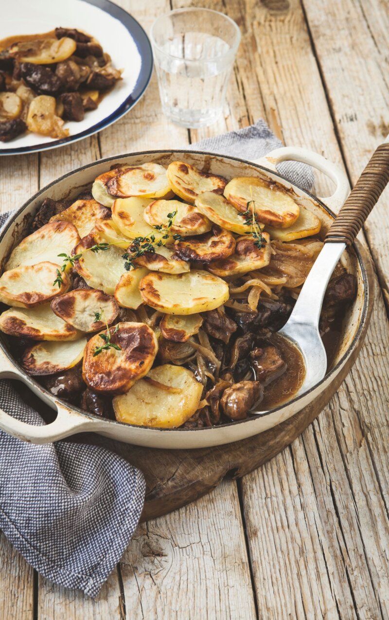 A rustic Lancashire hotpot of sliced potatoes baked atop a savoury meat and onion stew, inspired by the Hairy Bikers, served in a round ceramic pan with a serving spoon on a wooden table beside a blue serviette and glass of water.