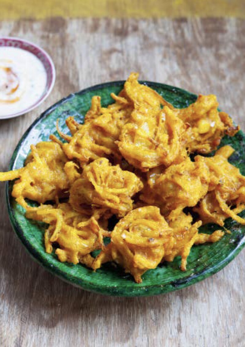 A green bowl of crispy, golden-brown pickled onion bhajis sits on a wooden table, reminiscent of a Hairy Bikers feast, with a small plate of dipping sauce in the background.