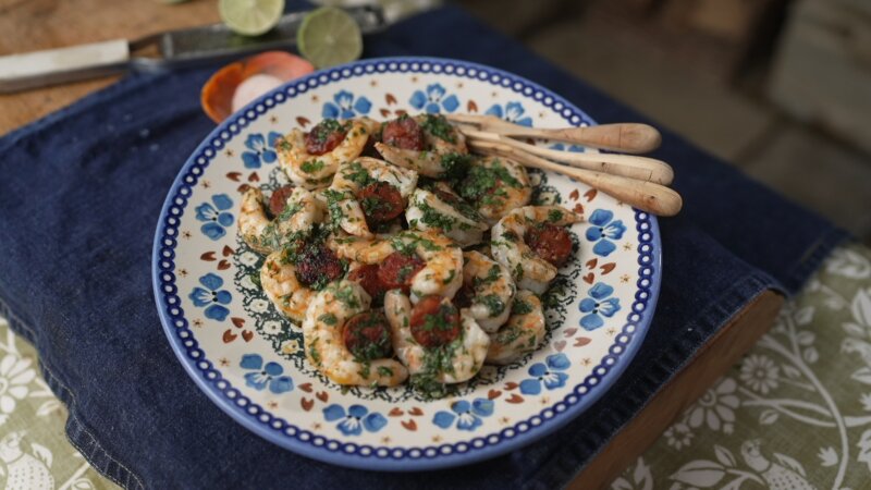 A ceramic plate with blue floral patterns showcases a shrimp dish garnished with herbs. Three wooden utensils rest on the plate's edge. The background includes lime slices, a knife, and Si King's Prawn and Chorizo bites, perfect for Simple for Summer moments.