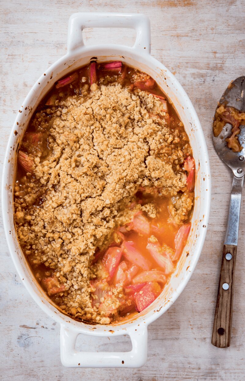An oval baking dish containing a rhubarb and orange crumble with visible rhubarb pieces and golden brown topping, alongside a used serving spoon.