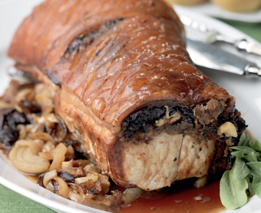 Roast Loin of Pork with crispy, golden-brown crackling rests on a plate, surrounded by caramelised onions and herbs—a classic Hairy Bikers recipe. A carving fork and knife are placed in the background.