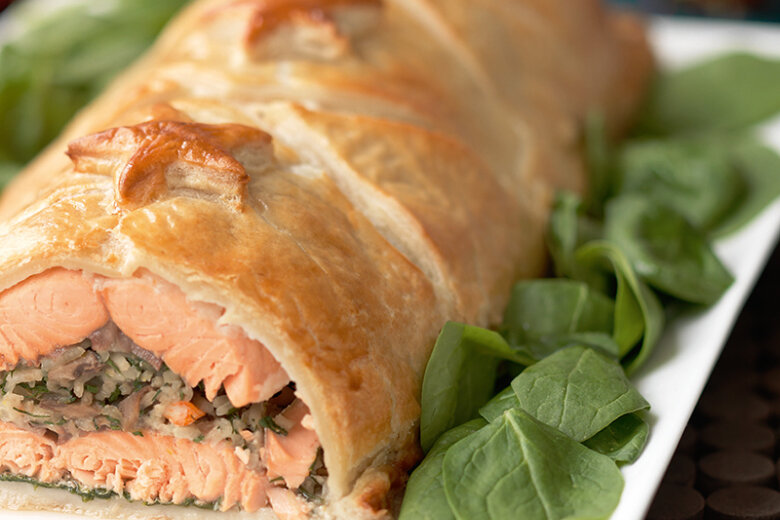 A close-up of Salmon Coulibiac, inspired by the Hairy Bikers’ Perfect Pies, with golden, flaky pastry and a pink salmon filling, sliced open and garnished with fresh spinach leaves on a white plate.