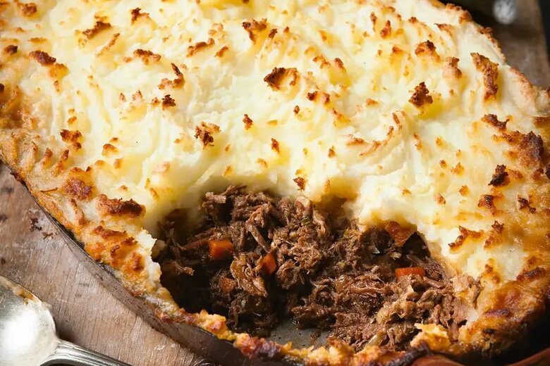 A delectable Shepherd's Pie, reminiscent of the Hairy Bikers' Best of comfort food, features a golden-brown mashed potato topping and a hearty filling of shredded beef and vegetables. A serving spoon and cloth lie ready beside this classic dish.