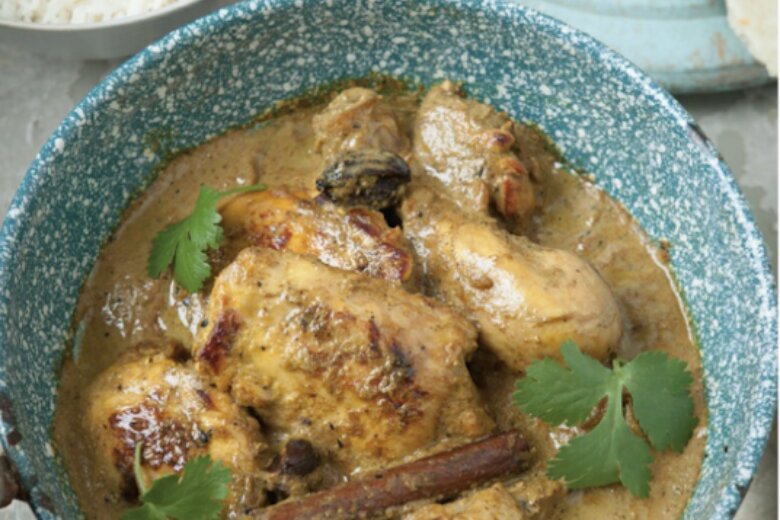 A blue bowl filled with Shorshe Murgi, a  curry garnished with coriander, accompanied by a side of white rice and flatbread in the background—a dish perfect for fans of 'Chicken & Egg cookbook by the Hairy Bikers.
