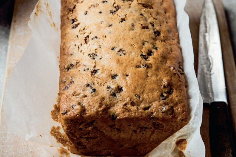 A spicy fruit loaf, from the Best of the Hairy Bikers' cookbook, rests in parchment paper on a wooden cutting board next to a bread knife.