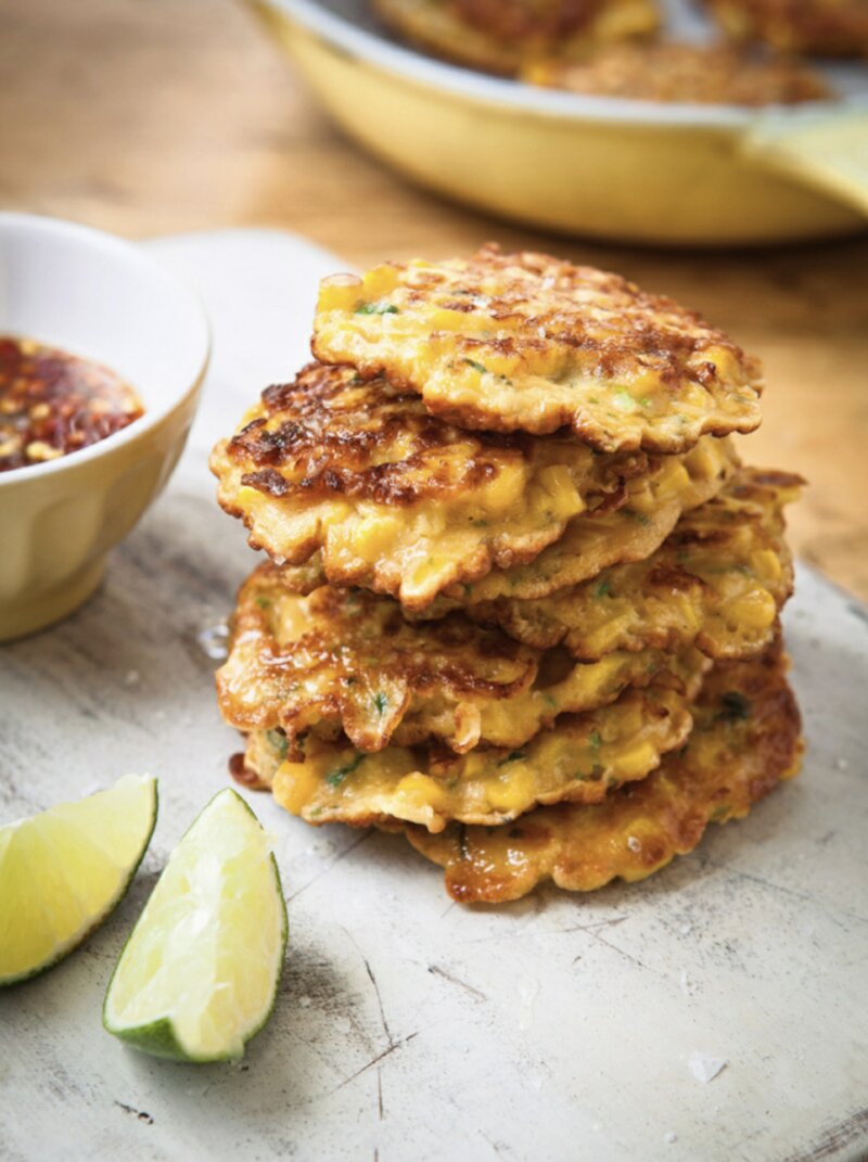 A stack of golden Sweetcorn Fritters from Hairy Dieters Fast Food sits on a cutting board, flanked by two lime wedges and a bowl of dipping sauce.