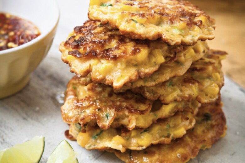 A stack of golden Sweetcorn Fritters from Hairy Dieters Fast Food sits on a cutting board, flanked by two lime wedges and a bowl of dipping sauce.