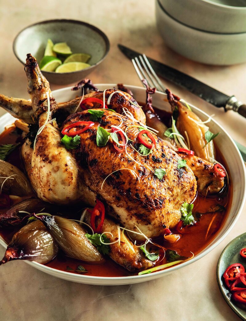 A Hairy Bikers Thai pot-roast chicken is garnished with herbs and sliced red chili, surrounded by whole onions and a rich sauce. Lime wedges rest on a small plate beside the dish, enhancing its vibrant flavours.