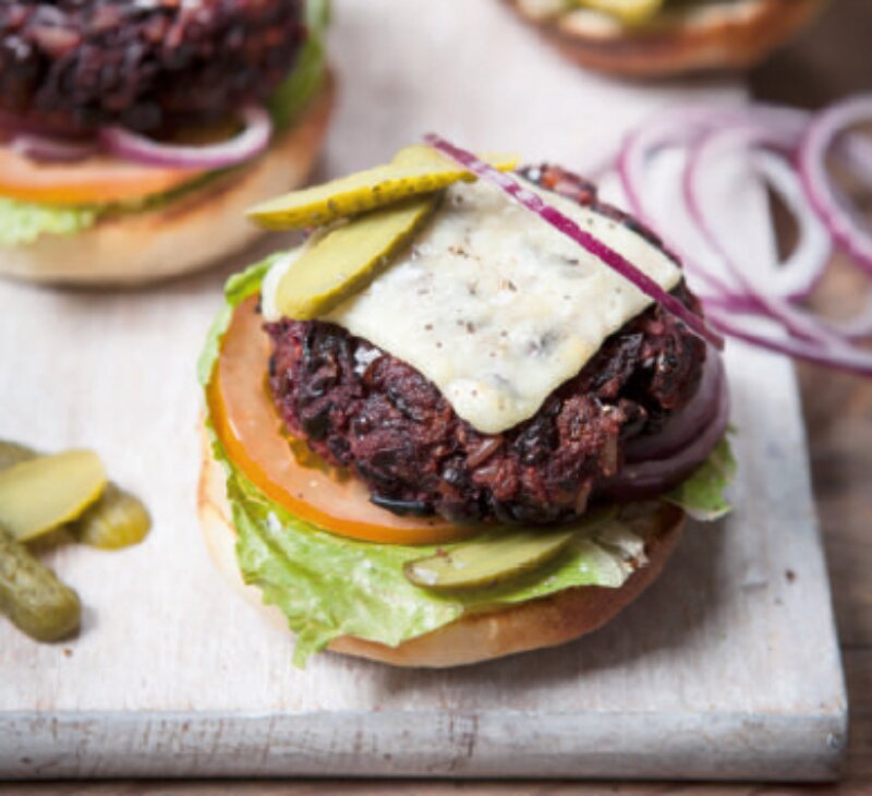 A Hairy Dieters Veggie Burger with a patty, melted cheese, lettuce, tomato slice, pickle slices, and red onion on a bun, partially assembled on a wooden surface.