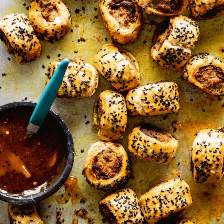 Golden, flaky Vindaloo Sausage Rolls from the Hairy Bikers Brilliant Bakes cookbook are topped with black sesame seeds and scattered on a baking tray beside a small bowl of dipping sauce with a green-handled spoon.
